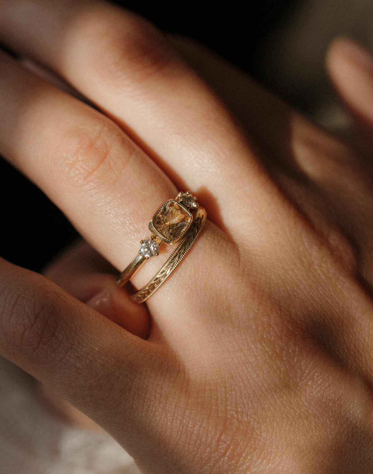 Close view of a hand in warm light wearing a yellow gold engagement ring with a cushion cut peach sapphire set east-west in a milgrain bezel and a round diamond on each side, stacked with a hand-engraved band edged in milgrain.