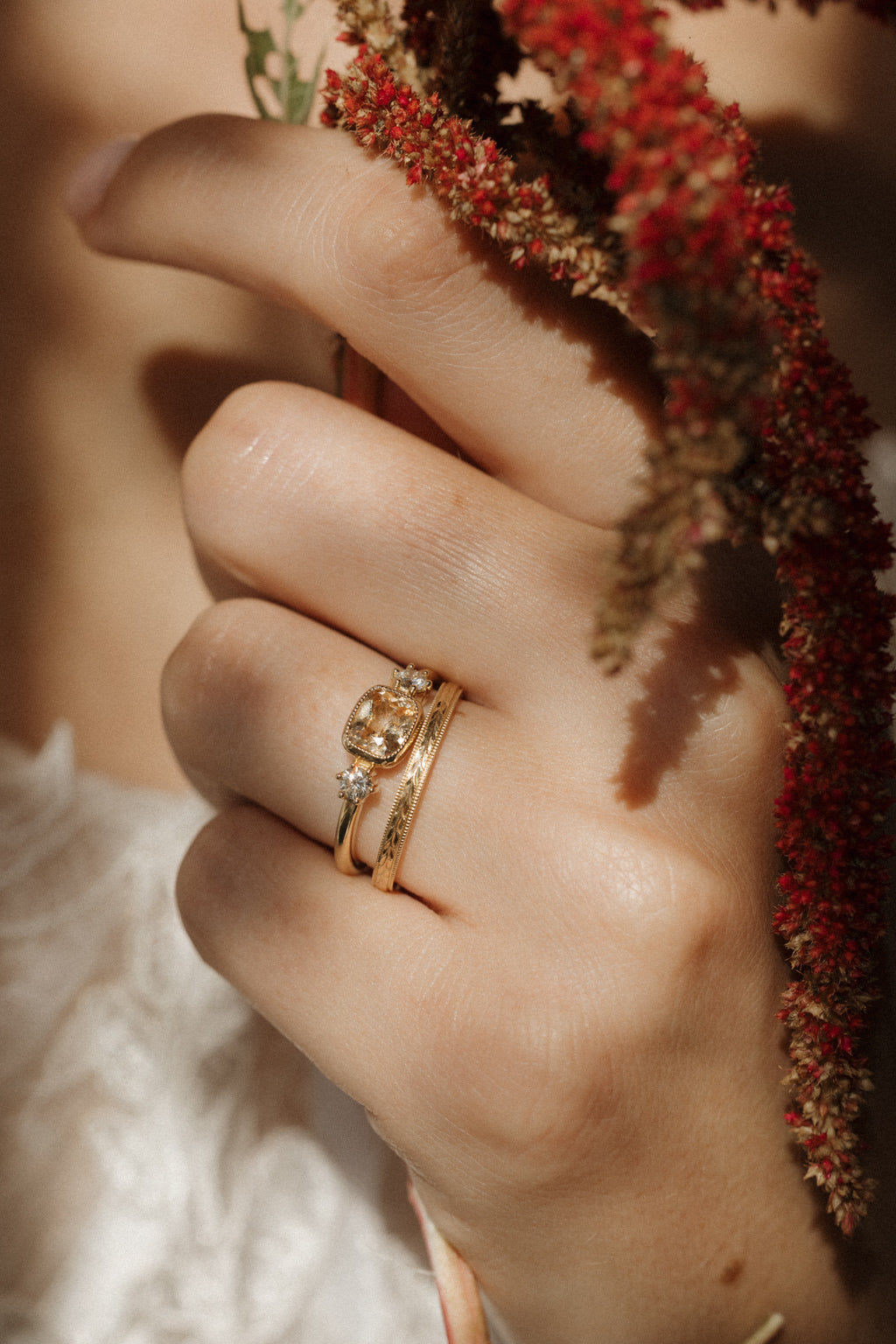 A hand holding stems of deep red amaranth, wearing a yellow gold peach sapphire engagement ring stacked with a hand-engraved wedding band, with a white lace sleeve below.