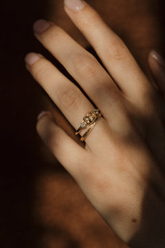 A hand in a streak of warm light against a dark background, wearing a yellow gold peach sapphire engagement ring stacked with a hand-engraved wedding band.
