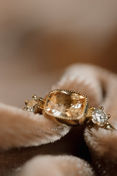 Close-up of a yellow gold ring nestled in folds of blush velvet, showing a cushion cut peach sapphire in a beaded milgrain bezel and a round diamond in prongs on either side.