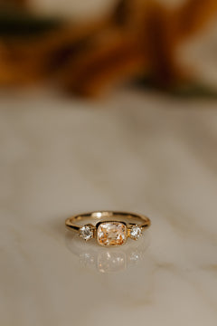 Front view of a yellow gold engagement ring with an east-west cushion cut peach sapphire in a milgrain bezel between two round diamonds, on a pale marble surface with its reflection below and warm blurred tones behind.