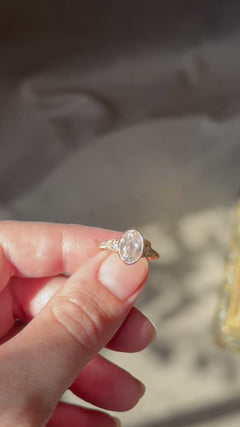 Video of fingers turning a yellow gold engagement ring in sunlight to show the bezel-set antique oval diamond, pear cut diamond side stones and wheat hand-engraved band from several angles, with an antique-style brass jewelry box blurred in the background.