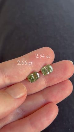 Video of two loose cushion cut green diamonds sitting side by side on the fingertips, shifting slightly in the light against a dark fabric background, with text overlays reading "2.66 ct" and "2.54 ct."