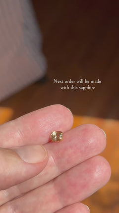 Video of a loose cushion cut peach sapphire held on a fingertip, shifting slightly as it catches warm sunlight against a blurred wood background, with the text overlay "Next order will be made with this sapphire."