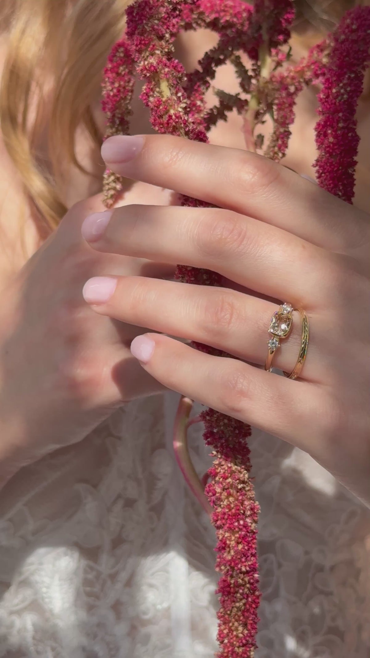 Close-up video of a hand holding red amaranth in front of the body, wearing a yellow gold engagement ring with an east-west cushion cut peach sapphire.
