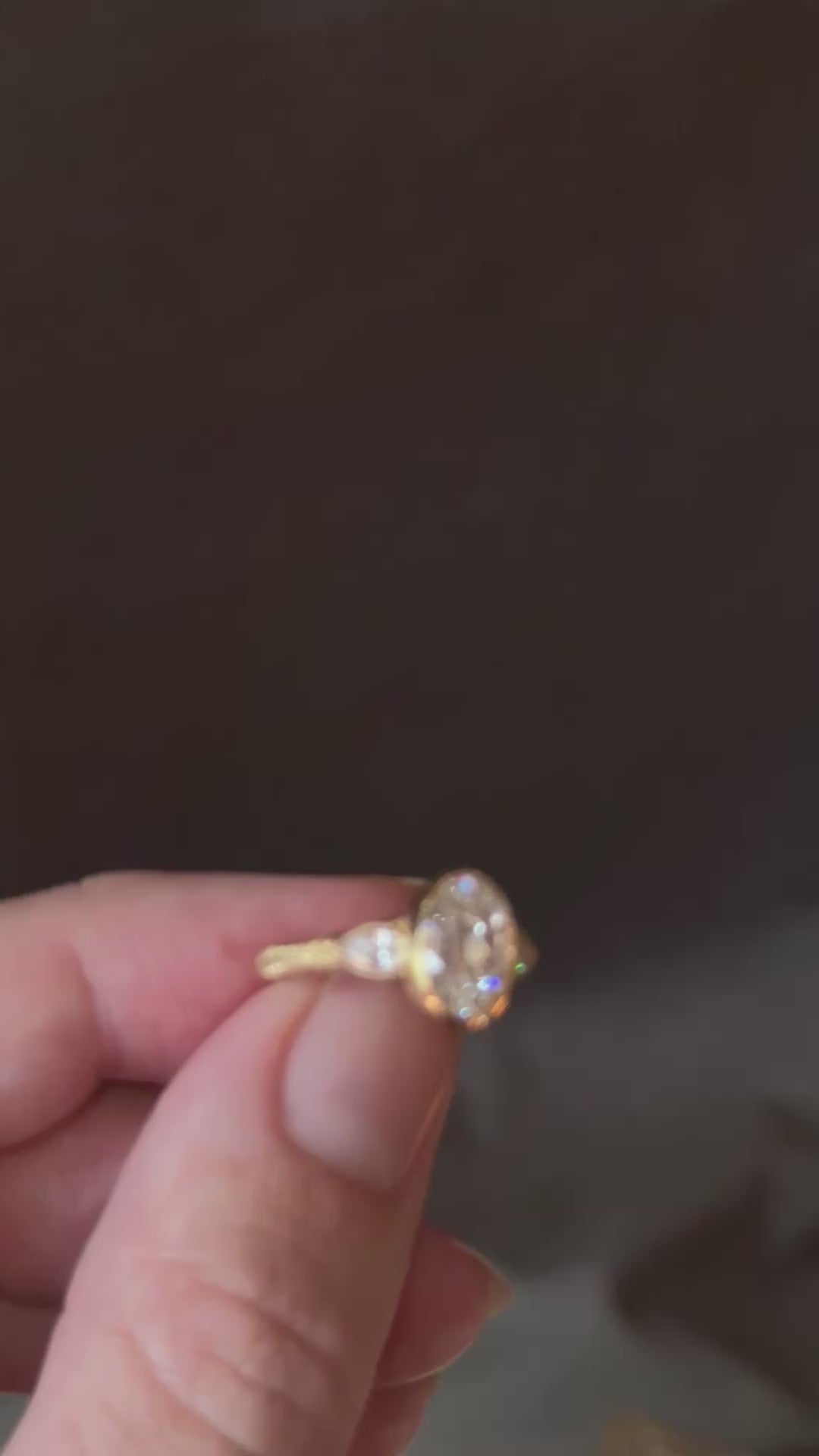 Video of fingers turning a yellow gold engagement ring against a dark background, showing the bezel-set antique oval diamond from the front and side, the pear cut diamond side stones, the star-set diamonds on the profile and the wheat hand-engraved band.
