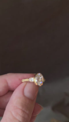 Video of fingers turning a yellow gold engagement ring against a dark background, showing the bezel-set antique oval diamond from the front and side, the pear cut diamond side stones, the star-set diamonds on the profile and the wheat hand-engraved band.