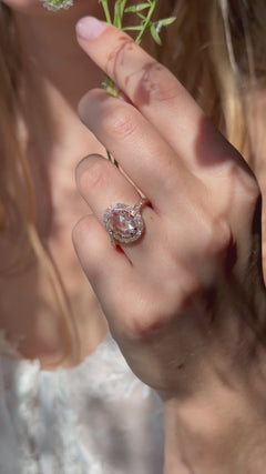 A close up video of a woman holding the stems of small white flowers in front of her partially visible face. On her hand she is wearing a rose gold and rose cut diamond engagement ring with four double prongs and a single cut diamond bypass halo.