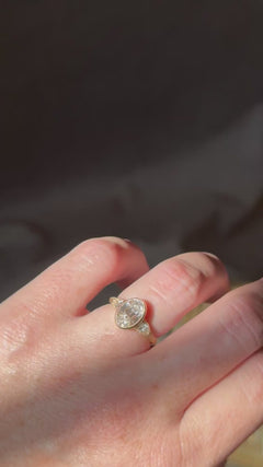 Video of a hand wearing a yellow gold engagement ring with a bezel-set antique oval diamond and pear cut diamond side stones, tilting slowly in warm sunlight against a dark background.