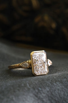 Angled front view of a yellow gold engagement ring with a radiant cut diamond in a bezel setting, flanked by pear cut diamond accents in milgrain-edged bezels on a wheat hand-engraved band, on dark gray velvet with an antique-style brass jewelry box blurred behind.