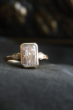 Front view of a yellow gold engagement ring with a radiant cut diamond in a bezel setting and pear cut diamond accents on a wheat hand-engraved band, on dark gray velvet with an antique-style brass jewelry box blurred behind.