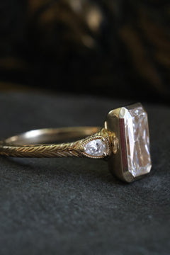 Angled side view of a yellow gold engagement ring with the bezel-set radiant cut diamond facing right, a pear cut diamond accent in a milgrain-edged bezel, and the wheat hand-engraved band, on dark gray velvet with an antique-style brass jewelry box blurred behind.