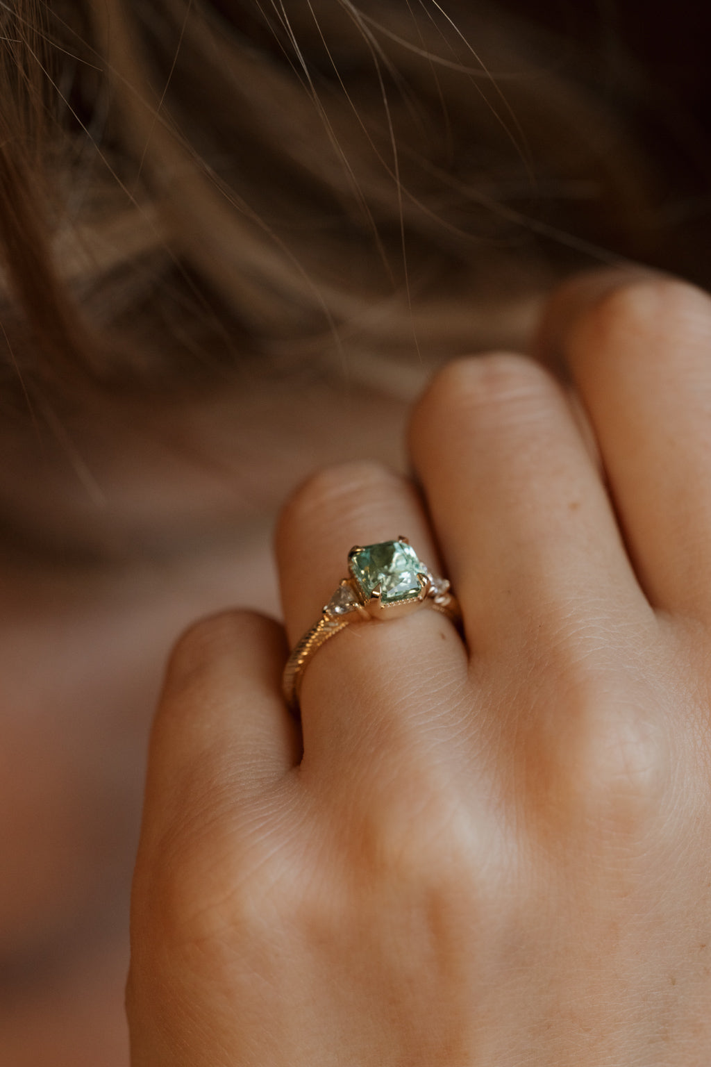 On-hand view of a yellow gold three stone engagement ring with a radiant cut green tourmaline in four corner claw prongs with milgrain detail, a trillion cut diamond on each side, and a hand-engraved band, the hand resting against loose light brown hair.