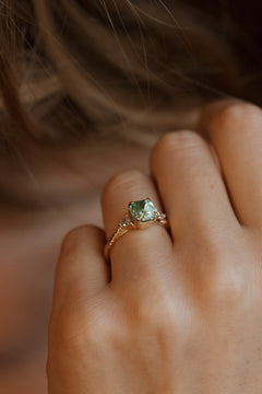 On-hand view of a yellow gold three stone engagement ring with a radiant cut green tourmaline in four corner claw prongs with milgrain detail, a trillion cut diamond on each side, and a hand-engraved band, the hand resting against loose light brown hair.