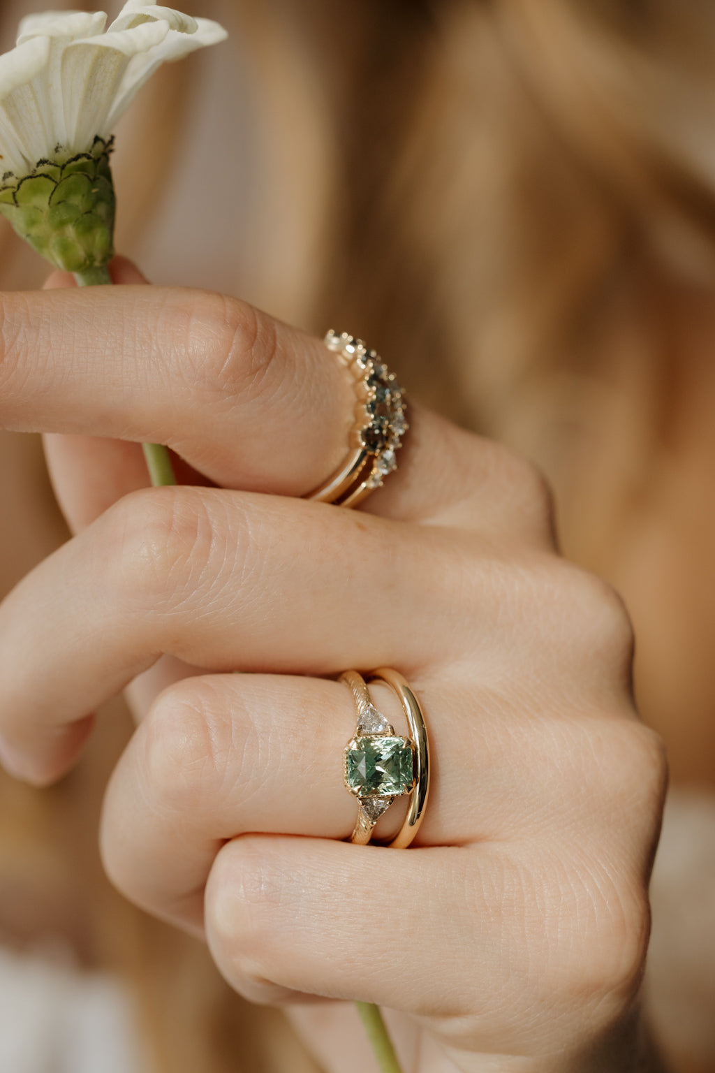 On-hand view of a yellow gold three stone engagement ring with a radiant cut green tourmaline and trillion cut side diamonds on a hand-engraved band, stacked with a plain yellow gold band, with two stacked bands set with light and dark stones on the index finger, the hand holding a white zinnia.