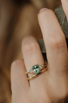 On-hand front view of a yellow gold three stone engagement ring with a radiant cut mint green tourmaline in corner claw prongs and a trillion cut diamond on each side, stacked with a plain yellow gold band, the hand holding a fold of olive woven fabric against a warm brown background.