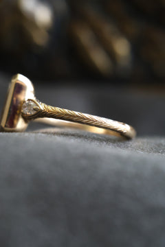 Close side profile of a yellow gold engagement ring showing the wheat hand-engraved band and a pear cut diamond accent in a milgrain-edged bezel beside the bezel-set radiant cut diamond, on dark gray velvet.