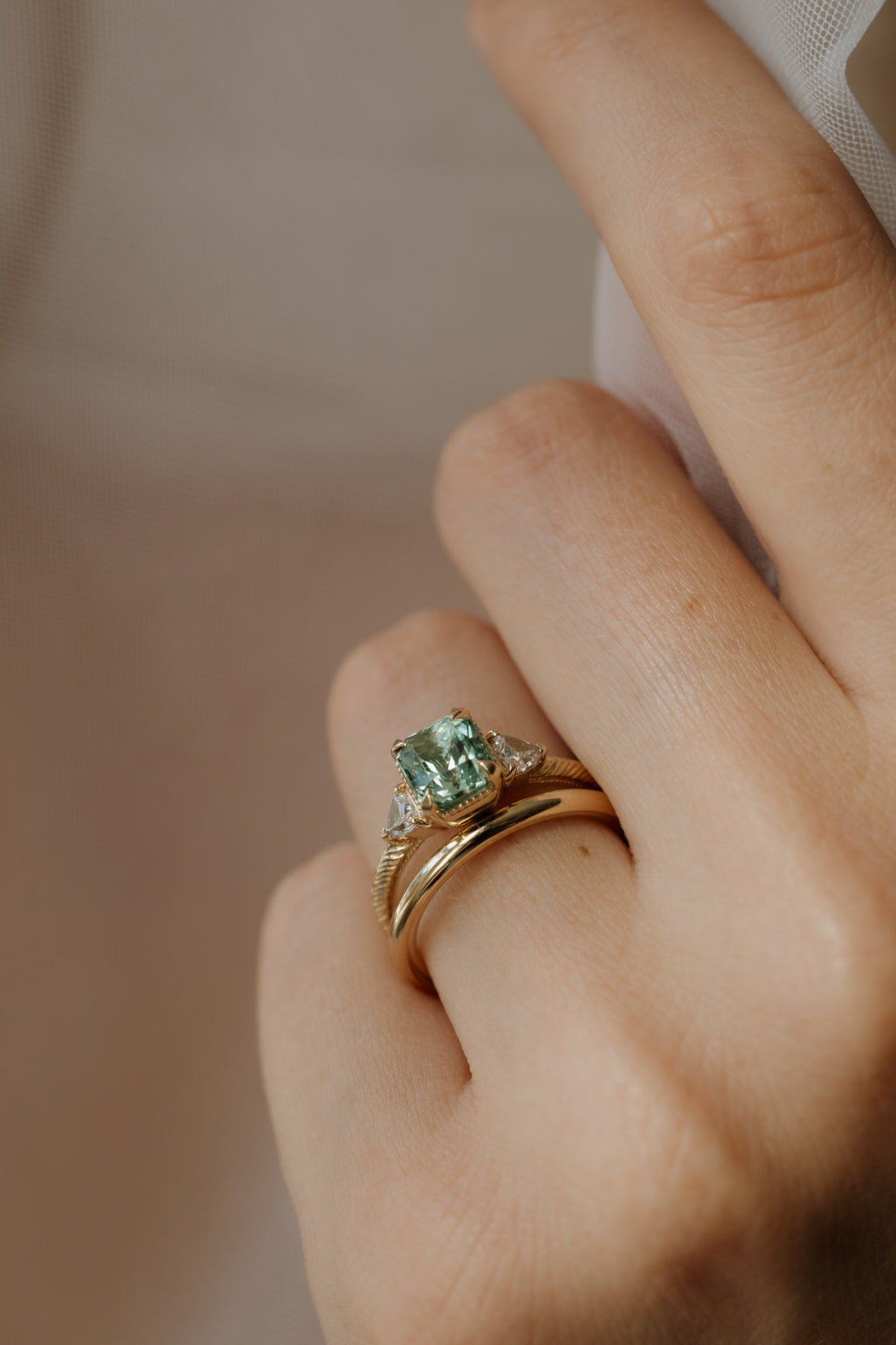 On-hand view of a yellow gold three stone engagement ring with a radiant cut green tourmaline in corner claw prongs, trillion cut side diamonds, and a hand-engraved band, stacked with a plain yellow gold band, the fingers holding sheer white tulle.
