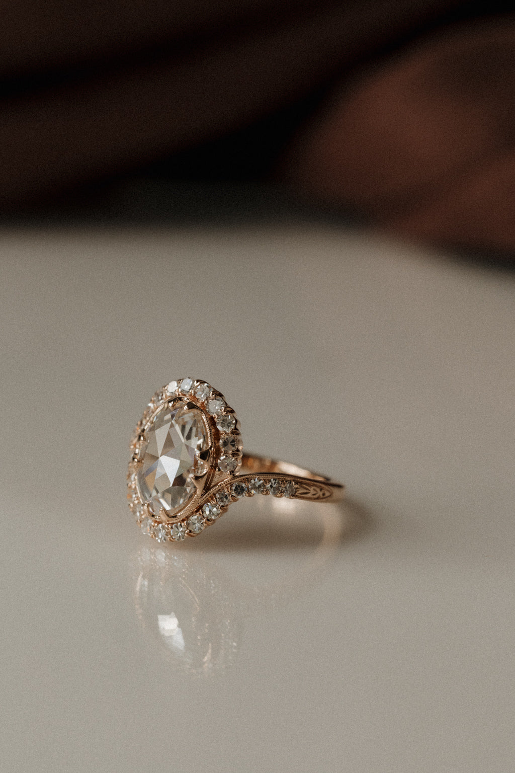A rose gold engagement ring from a side view, sitting on a white surface. The ring has an oval rose cut diamond center stone with four double prongs in a compass setting. The band has a small engraving on either side where a single cut diamond bypass halo starts