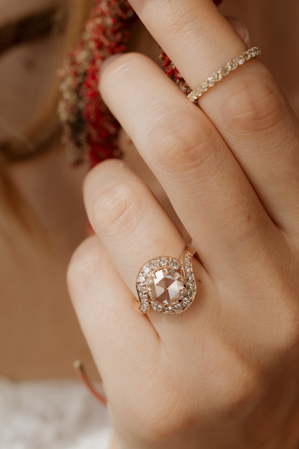 Close up photo of a hand wearing an engagement ring on the ring finger and a thin diamond eternity band on the index finger, with blonde hair and dried red amaranth out of focus behind. The ring is a rose gold curved bypass setting with a round rose cut diamond in a halo of single cut diamonds.