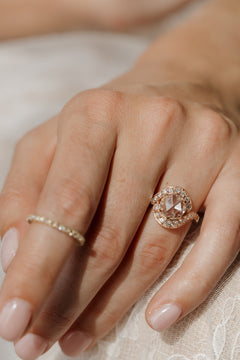 Close up photo of a hand resting on pale cream fabric, wearing an engagement ring on the ring finger and a thin diamond eternity band on the index finger. The ring is a rose gold curved bypass setting. It has a round rose cut diamond surrounded by a halo of single cut diamonds with milgrain along the edges.