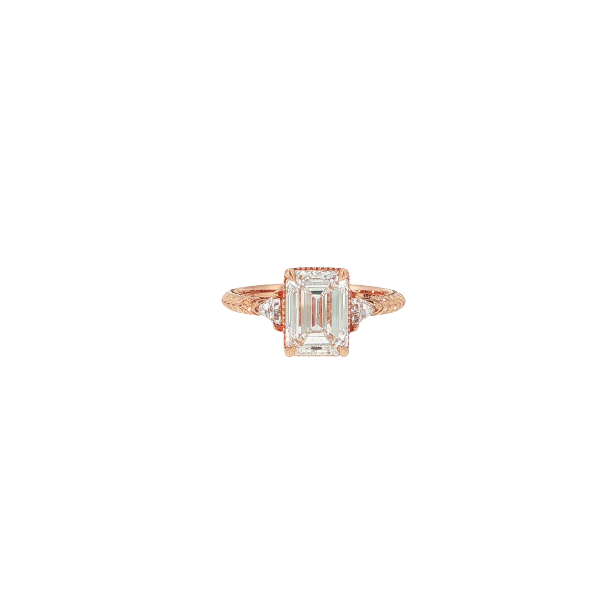 Close-up of a three stone engagement ring featuring an emerald cut diamond with triangle cut accent diamonds, set in rose gold with a hand-engraved band and milgrain detail.