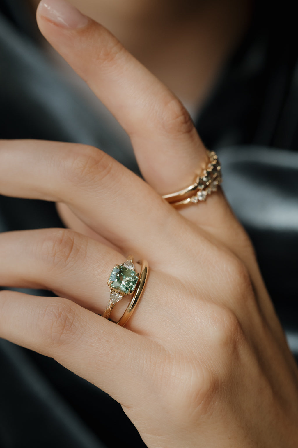 On-hand view of a yellow gold three stone engagement ring with a radiant cut green tourmaline, trillion cut side diamonds, and a hand-engraved band, stacked with a plain yellow gold band, with two stone-set bands on the index finger, against dark gray satin.
