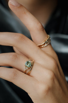 On-hand view of a yellow gold three stone engagement ring with a radiant cut green tourmaline, trillion cut side diamonds, and a hand-engraved band, stacked with a plain yellow gold band, with two stone-set bands on the index finger, against dark gray satin.