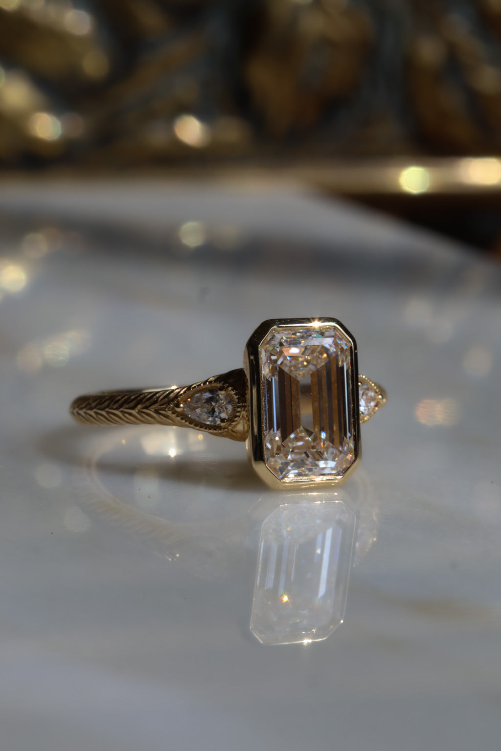 Close up side profile of a bezel emerald cut diamond three stone engagement ring with pear cut diamond accent stones on either side with milgrain details, the band is yellow gold and hand engraved.