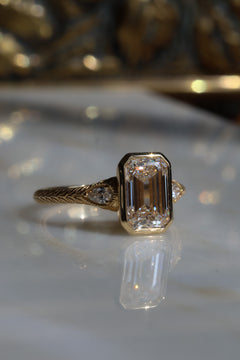 Close up side profile of a bezel emerald cut diamond three stone engagement ring with pear cut diamond accent stones on either side with milgrain details, the band is yellow gold and hand engraved.