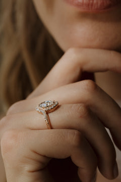 A close up of a woman with her hand under her chin, she is wearing a rose gold engagement ring on her hand. The engagement ring has a double prong set oval rose cut diamond with a single cut bypass halo surrounding it. 