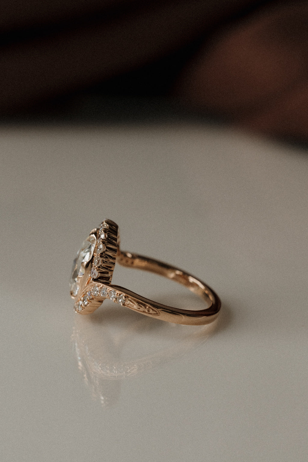 Side profile view of a rose gold engagement ring resting on a white surface. The ring has an oval shaped rose cut center stone, and a bypass halo made of twenty-eight single cut diamonds. Where the band and the halo meet there is a small engraving.