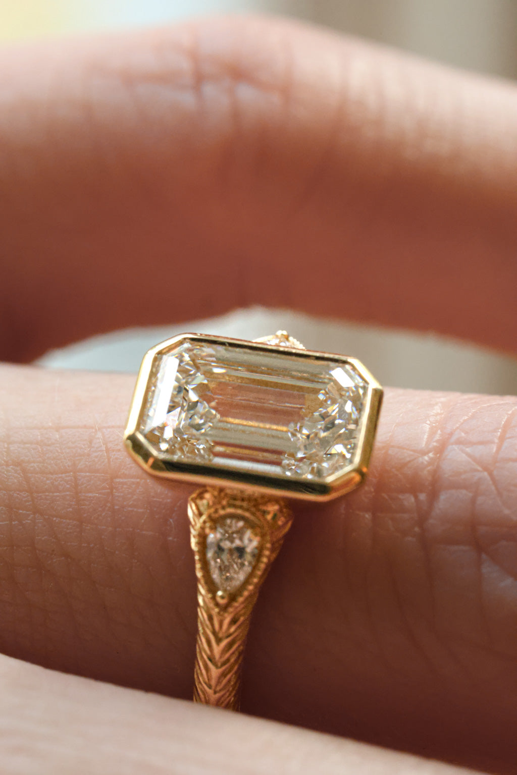 A close up side view of a finger wearing a bezel set emerald cut diamond engagement ring. The ring is yellow gold, hand engraved, and has two pear shaped side stones with a milgrain border.