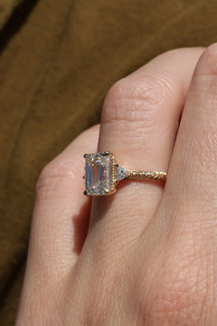 Close up photo of a Moonryvr engagement ring on a finger tilted to the side. The ring is a yellow gold hand-engraved band with milgrain details along the side of the band. It has an emerald cut diamond in a three stone setting with four claw prongs around the center stone, and the accent diamonds are triangle cut.