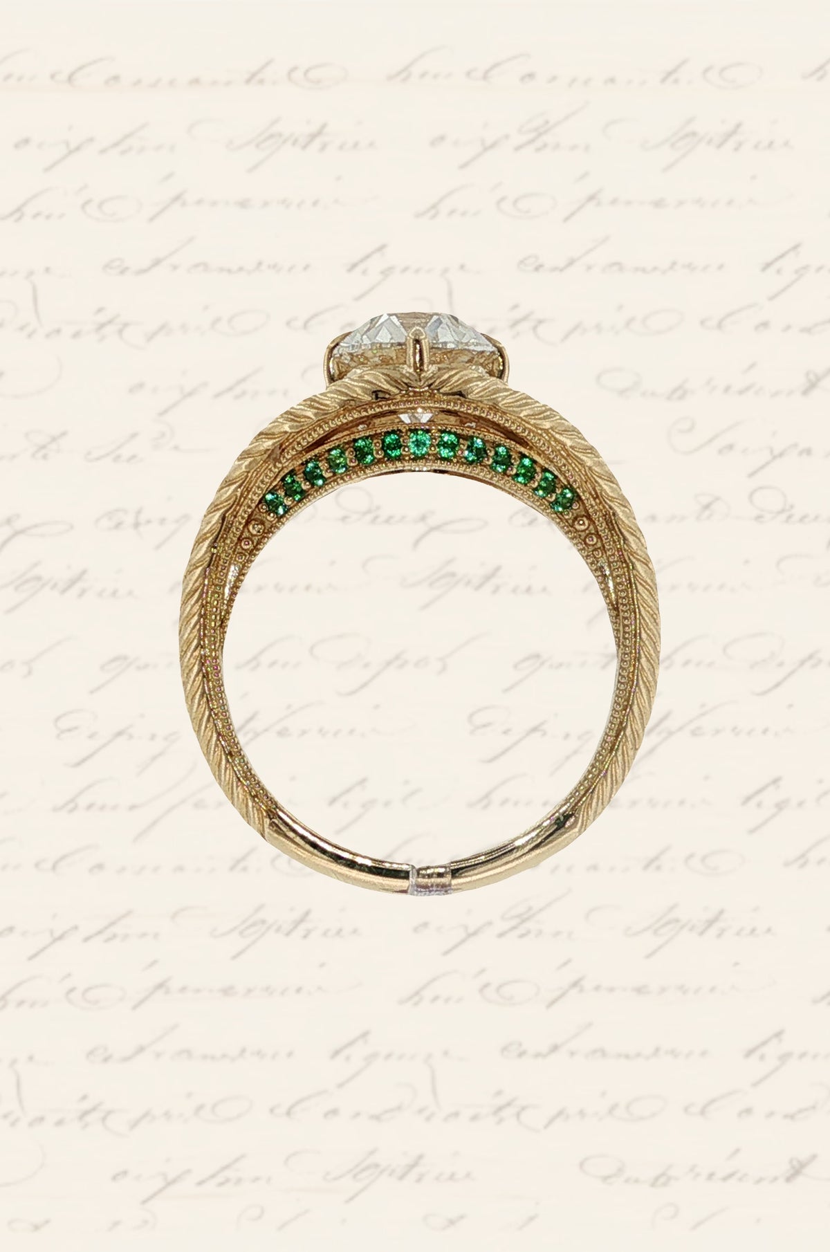 Side view of a Moonryvr engagement ring with a yellow gold, old-world, hand engraved, split shank band. The center stone is an old mine cut diamond with four claw prongs in a compass setting. The split shank has a hidden halo made of emeralds on the lower profile of the side view. 