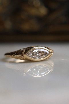 Close up photo of an engagement ring at a three quarter angle on a white reflective surface, with a dark blurred background. The ring is a chunky yellow gold band with fine engraved lines along the shoulders. It has a marquise cut diamond set east-west in a bezel, reflected in the surface below.