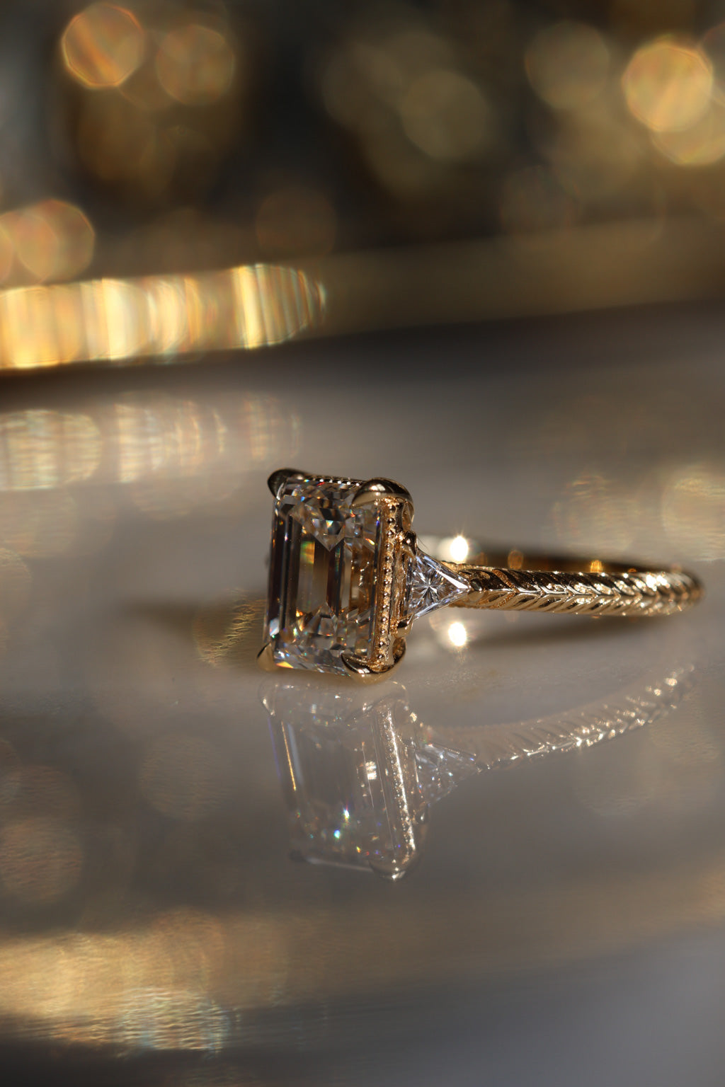 A close up side view picture of an engagement ring resting on a marble surface with a soft blur around it. The ring is a yellow gold hand-engraved band and milgrain details. It is a three stone setting with an emerald cut diamond set with four claw prongs and triangle accent diamonds.  