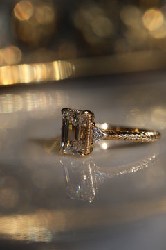 A close up side view picture of an engagement ring resting on a marble surface with a soft blur around it. The ring is a yellow gold hand-engraved band and milgrain details. It is a three stone setting with an emerald cut diamond set with four claw prongs and triangle accent diamonds.  