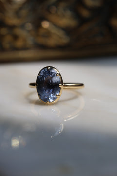 Close up side profile photo of an engagement ring standing upright on a beige reflective surface. The ring is a thin, fully rounded yellow gold band. The oval blue Ceylon sapphire sits in a collet cup that curves smoothly into the band.
