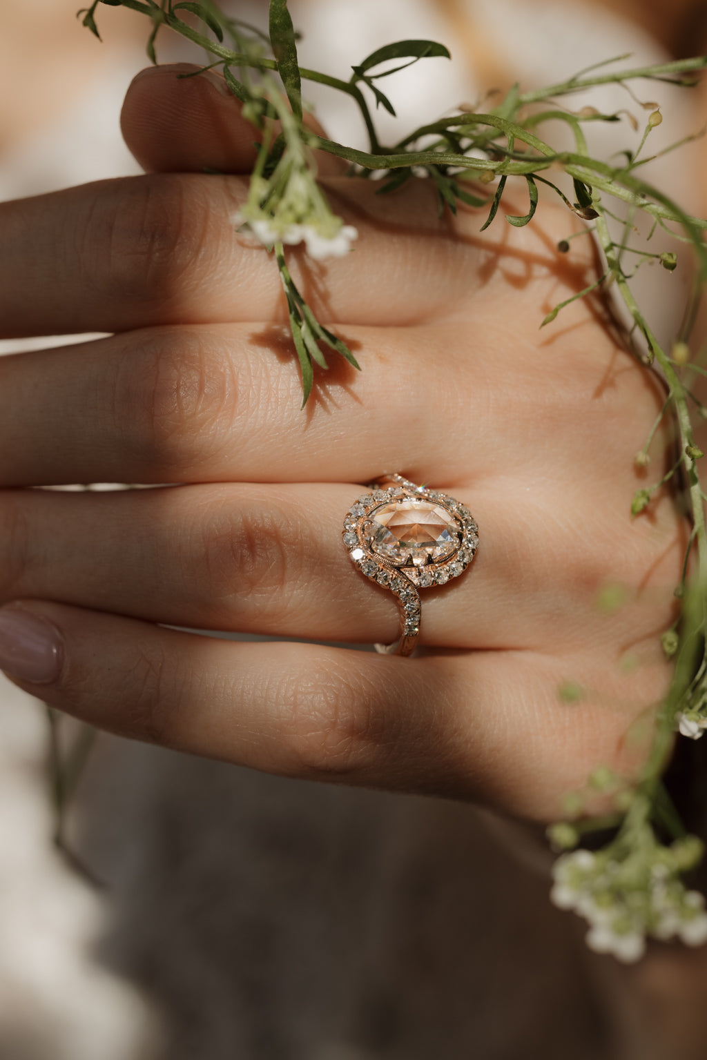 A hand wearing a rose gold engagement ring with small white flowers over it. The engagement ring is a double prong rose cut diamond with a  bypass single cut diamond halo. The band has small engraved details where the halo ends. 