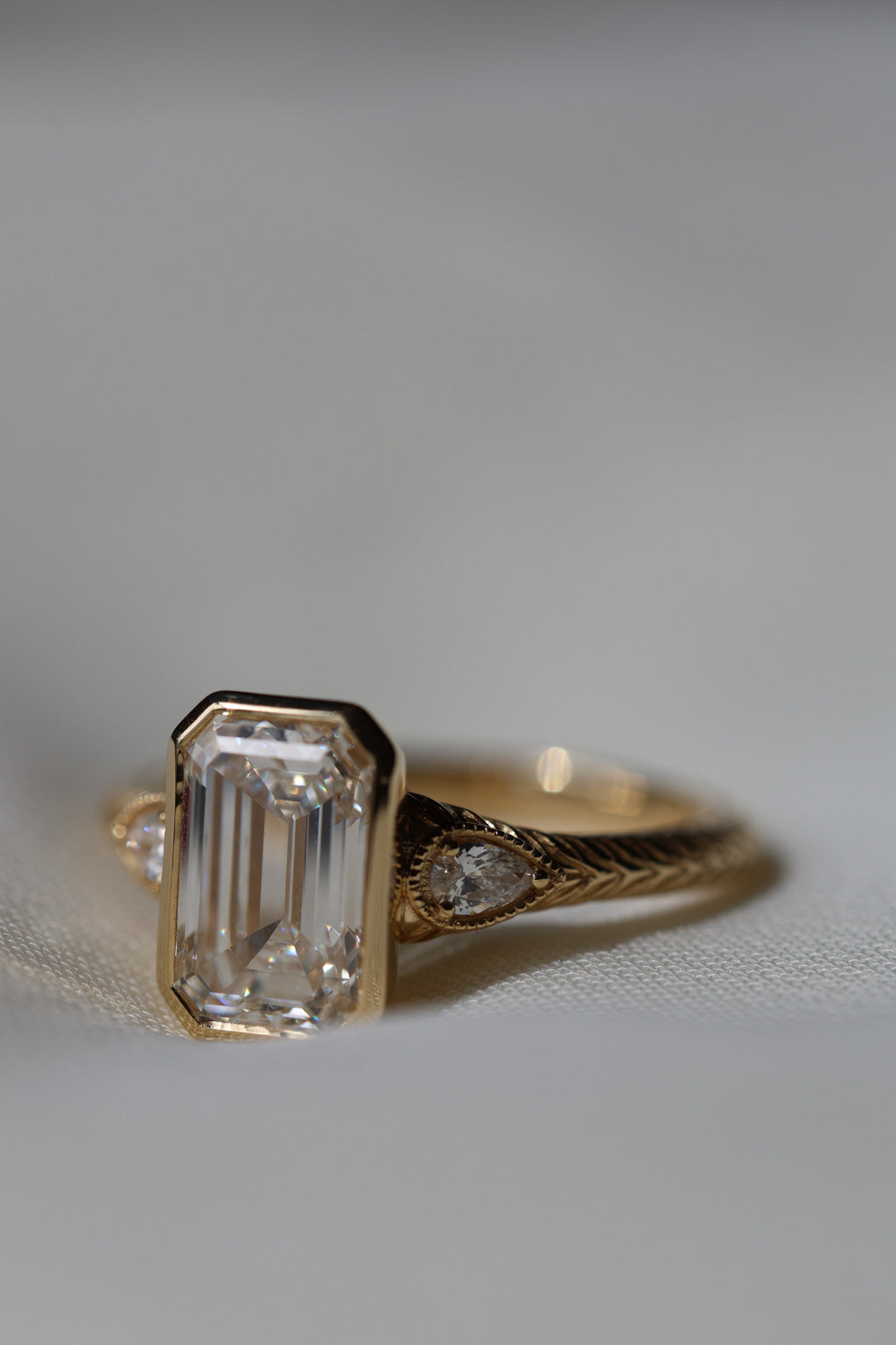 A close up side view of a hand engraved yellow gold three stone engagement ring. The center stone is a bezel set emerald cut diamond, two pear cut accent diamonds are on either side with a milgrain edge.