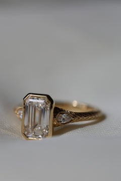 A  close up of a yellow gold hand engraved engagement ring sits on white fabric. turned to a slight side view angle. The ring has an emerald cut diamond in a bezel setting with two pear cut side stones with a milgrain border.