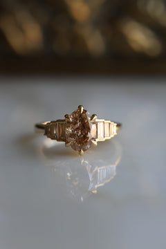 Front view of a desert diamond engagement ring, a fancy light brown color, with a six claw prong setting. There are four baguette cut white diamonds on either side of the center stone, set in a descending height in a stair step fashion. The side stones are bezel set, the ring is yellow gold. 