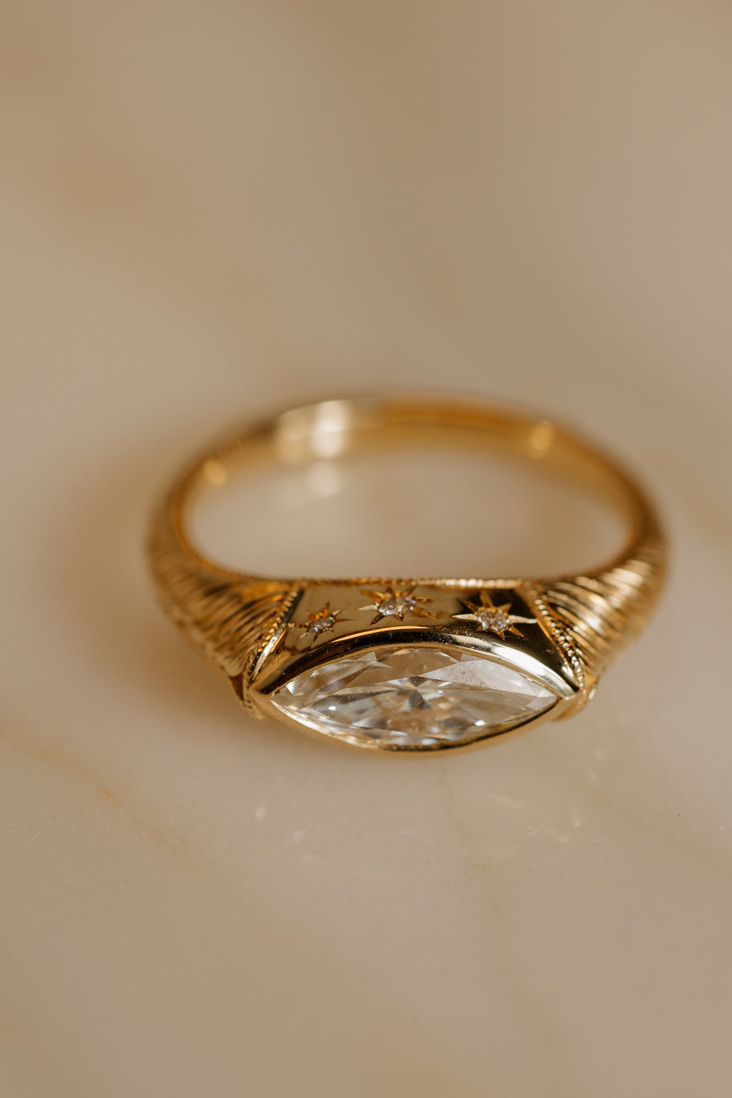 Close up photo of an engagement ring on a warm cream surface, angled to show the top of the setting. The ring is a chunky yellow gold band with fine engraved lines along the shoulders. It has a marquise cut diamond set east-west in a bezel, with small star-set diamonds worked into the gold above the stone.