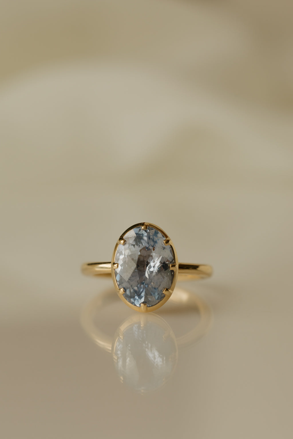 Close up photo of an engagement ring on a white reflective surface. The ring is a thin yellow gold band. It has an oval blue Ceylon sapphire held in a collet setting with eight prongs, and the stone's facets show both pale and deeper blue.