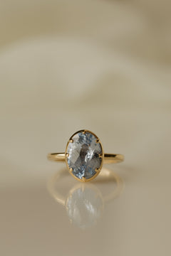 Close up photo of an engagement ring on a white reflective surface. The ring is a thin yellow gold band. It has an oval blue Ceylon sapphire held in a collet setting with eight prongs, and the stone's facets show both pale and deeper blue.