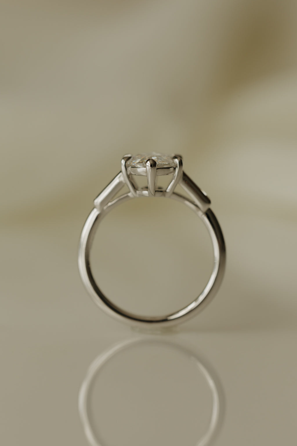 End-on profile view of a white gold engagement ring standing upright, showing the round band, the claw prongs lifting the old mine cut oval diamond, and a tapered baguette on each shoulder, on a reflective cream surface.